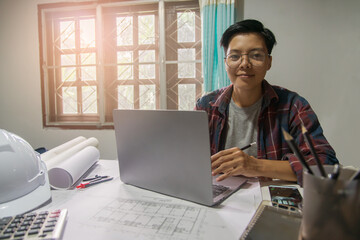 Concept people, smiling Asian engineer on the desk with a blueprint in the home office, Sunset light.Selective Focus