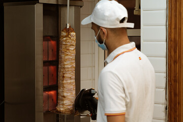 Uniformed chef, cutting and preparing chicken and beef doner kebabs using a prevention face mask at...