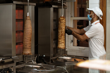 Uniformed chef, cutting and preparing chicken and beef doner kebabs using a prevention face mask at a fast food stand.