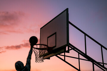 silhouette alone dunking man at sunset. basketball player scoring. Young silhouetted man jumping high to the target basketball hoop achieves to score with a flying slam dunk at sunset. © William