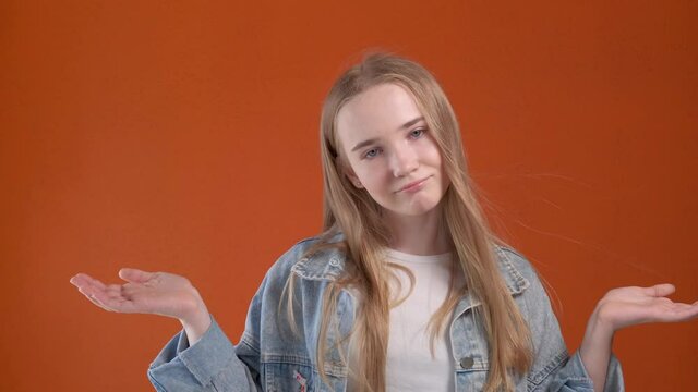 The blonde girl is at a loss and spreads her arms to the sides. Human emotions. Orange background.