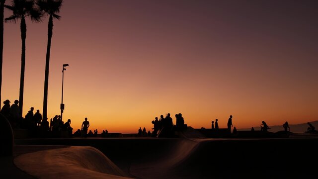 Silhouette of young jumping skateboarder riding longboard, summer sunset background. Venice Ocean Beach skatepark, Los Angeles California. Teens on skateboard ramp, extreme park. Group of teenagers.