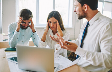 Fototapeta premium Tired office workers feeling headache during deadline reading email with bad news from colleague about project, unhappy male and female partners near desktop with blueprint and modern laptop computer
