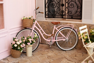 Fototapeta na wymiar Vintage bike with basket with bouquet flowers of roses stands near cafe of bakery. Eco transport. pink bicycle with flower pots with roses stands on street coffee shop. wedding decorations. 