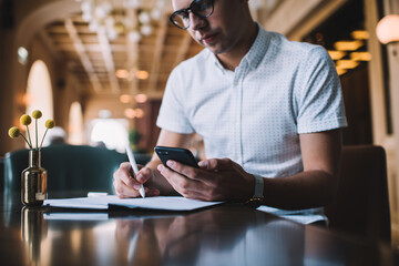 Crop male freelancer messaging with partner using smartphone and notepad in modern cafe