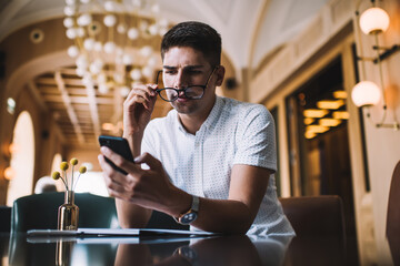 Pensive young man taking off eyeglasses while using smartphone in cafe