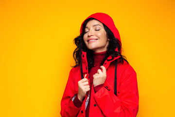 Delighted positive brunette woman smiling, wear turtleneck and red raincoat with hood, enjoys the rainy weather, has good mood, closed eyes, isolated on studio yellow background. Autumn season.