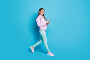 Fototapeta na wymiar Full size profile photo of attractive lady blogger hold telephone walk street browsing phone addicted user wear casual violet jacket sweater trousers shoes isolated blue color background