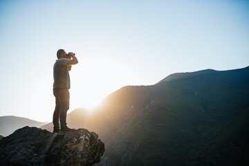 young man looking through binoculars on a mountain at sunset. Travel concept