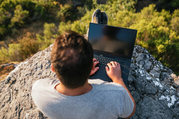 Young man sitting on a rock overlooking the mountain working with a laptop. Teleworking concept