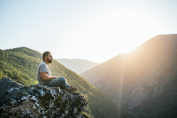 Fototapeta premium Young man sitting on a rock overlooking the mountain working with a laptop. Teleworking concept