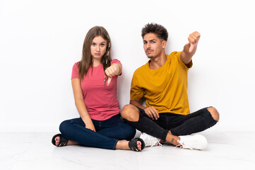 Obraz na płótnie Canvas Young couple sitting on the floor isolated on white background showing thumb down with negative expression