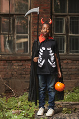 Vertical full shot portrait of cool African American boy dressed up as devil with red horns and scythe standing against old building looking at camera