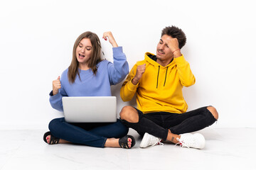 Young couple with a laptop sitting on the floor isolated on white background celebrating a victory