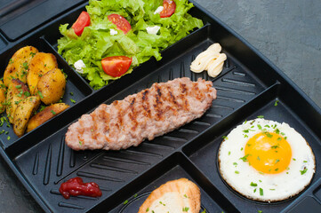 Restaurant lunch, content for the site. Breakfast, cutlet or steak, tomato salad, fried potatoes, croutons and fried egg on black plate.