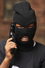 Vertical close-up head and shoulders portrait of cool little boy dressed up as gangster for Halloween wearing balaclava holding toy gun smiling at camera