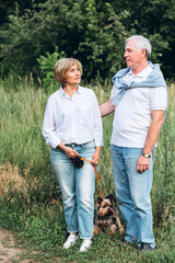 A mature couple is walking with a dog in a park. Elderly couple resting in nature with a dog. Close-up portrait of an elderly man and woman in white shirts and jeans. Stylish and modern grandparents.