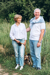 A mature couple is walking with a dog in a park. Elderly couple resting in nature with a dog. Close-up portrait of an elderly man and woman in white shirts and jeans. Stylish and modern grandparents.