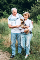 A mature couple is walking with a dog in a park. Elderly couple resting in nature with a dog. Close-up portrait of an elderly man and woman in white shirts and jeans. Stylish and modern grandparents.