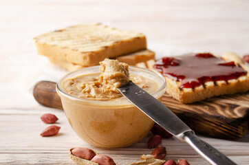 Low angle view at glass bowl with peanut butter on white wooden table with toasts and jam aside....