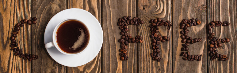 top view of cup of coffee on saucer and coffee lettering made of beans on wooden surface, panoramic shot