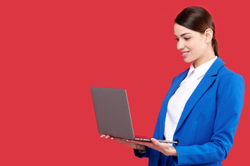 Young businesswoman in blue suit holding laptop and smiling on red isolated background