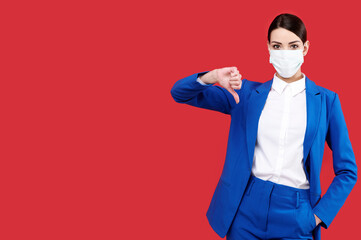 Caucasian business woman wearing protective medical mask and blue costume showing thumb down on red isolated background