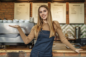 Nice stylish cheerful and attractive blonde girl, barista in casual shirt and apron, holding hand near bar counter