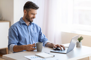 Fototapeta premium Home Office. Smiling Freelancer Man Checking Emails On Laptop And Drinking Coffee
