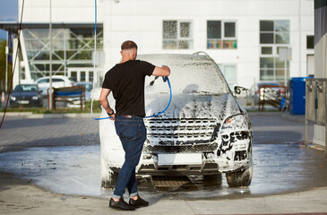 Back view of a sporty man holding a hose with soap and washing his car outdoors, white foam on a black car