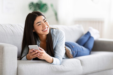 Carefree Asian Girl Enjoying Rest On Comfortable Sofa With Smartphone, Sincerely Smiling