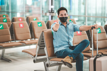 Traveler Asian man with luggage wearing mask protect from coronavirus sitting on social distancing chair showing thumb up with confidence