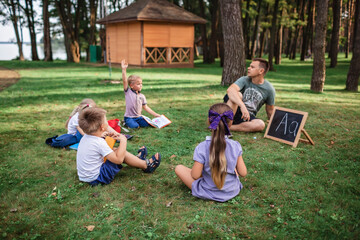 Back to school. Kindergarten and elementary scholars sitting with teacher on grass at open-air class