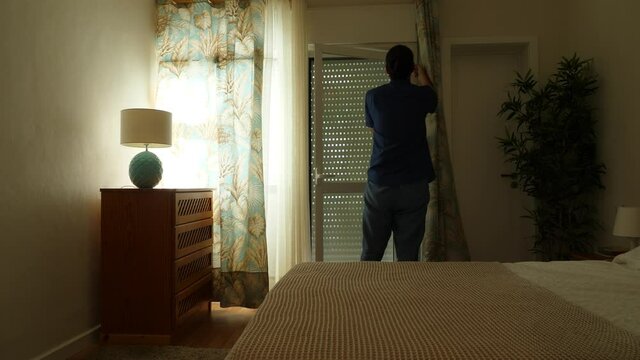 Man raise up window and door roller shutters, walk out to small balcony. Little bedroom become lit by bright daylight. Made bed and simple furniture seen in room