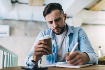 Serious man making notes in planner while drinking coffee