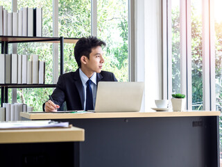 Fototapeta premium Young Asian businessman in suit working with laptop computer in office with green tree background outside.