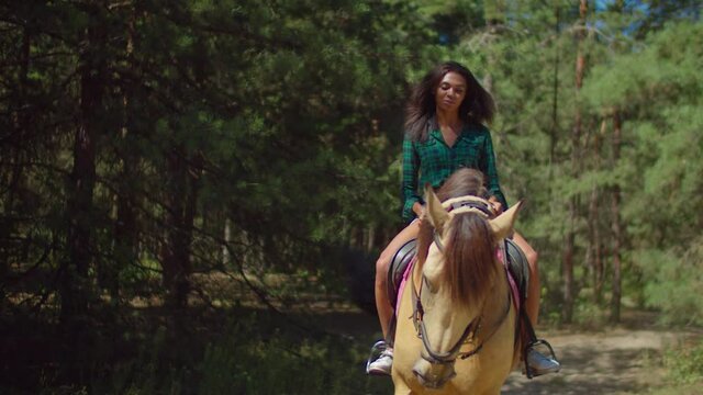 Positive Attractive Young African American Woman In Casual Clothes Enjoying Horseback Riding On Brown Purebred Horse In Forest During Summer Vacations In Countryside, Expressing Happiness And Joy.