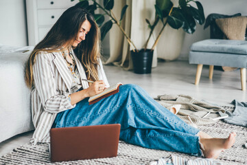 Elegant young woman in blue denim jeans writes in paper notebook sitting on floor near laptop at...