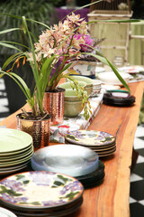 Orchids on a wooden table in a copper vase, surrounded by plates.