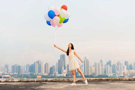 Happy girl jumping with colorful balloons on rooftop balcony with cityscape. Portrait of funny girl against sunset sky background. Active girl having fun on summer vacation. Freedom concept - Powered by Adobe
