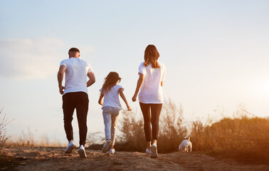 Back view of sporty and beautiful family of three jogging with their jack russell terrier outside the city on the village road on the sunset