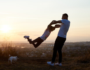 Silhouette of a father swinging his daughter by the arms on the hill on the setting sun in the...