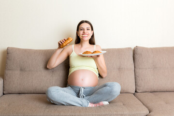 Fototapeta premium Young pregnant woman enjoys eating croissant on the sofa. Unhealthy pastry during pregnancy concept