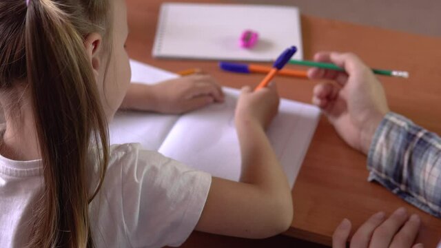 Preschool learning.A little girl with a teacher sits at a school desk and draws in a notebook with a pen.Drawing.4k