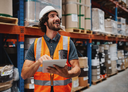 Waist up portrait of young man wearing reflective jacket holding digital tablet standing in factory warehouse smiling