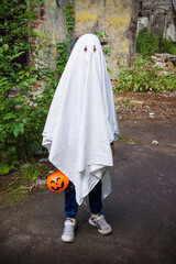 Child in halloween ghost costume with a basket of pumpkin on the background of broken old wall and trees