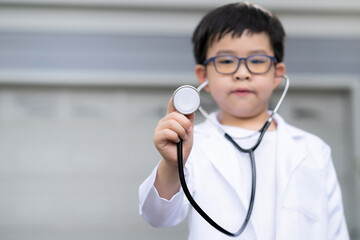 Asia boy wearing a medical suit and holding a doctor stethoscope on his hand standing at home. Feeling happy and confident. Focus on the stethoscope. Education and medical concept