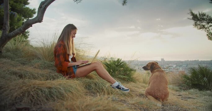 Pleasant young female working using laptop smiling with lovely dog in park outdoors, trendy girl in red caged shirt with computer online in social networks chatting with friends with animal