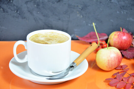White cup of coffee with cappuccino cream on an orange background with autumn leaves, apples and red berries. Fall season, leisure time and coffee break concept. Autumn leaves and hot steaming coffee