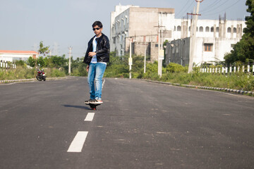 Smart Sporty Young Indian Boy In Black Jacket And Blue Jeans On Skateboard in Action Skating Along White Line Marks On Black Asphalt Concrete Road